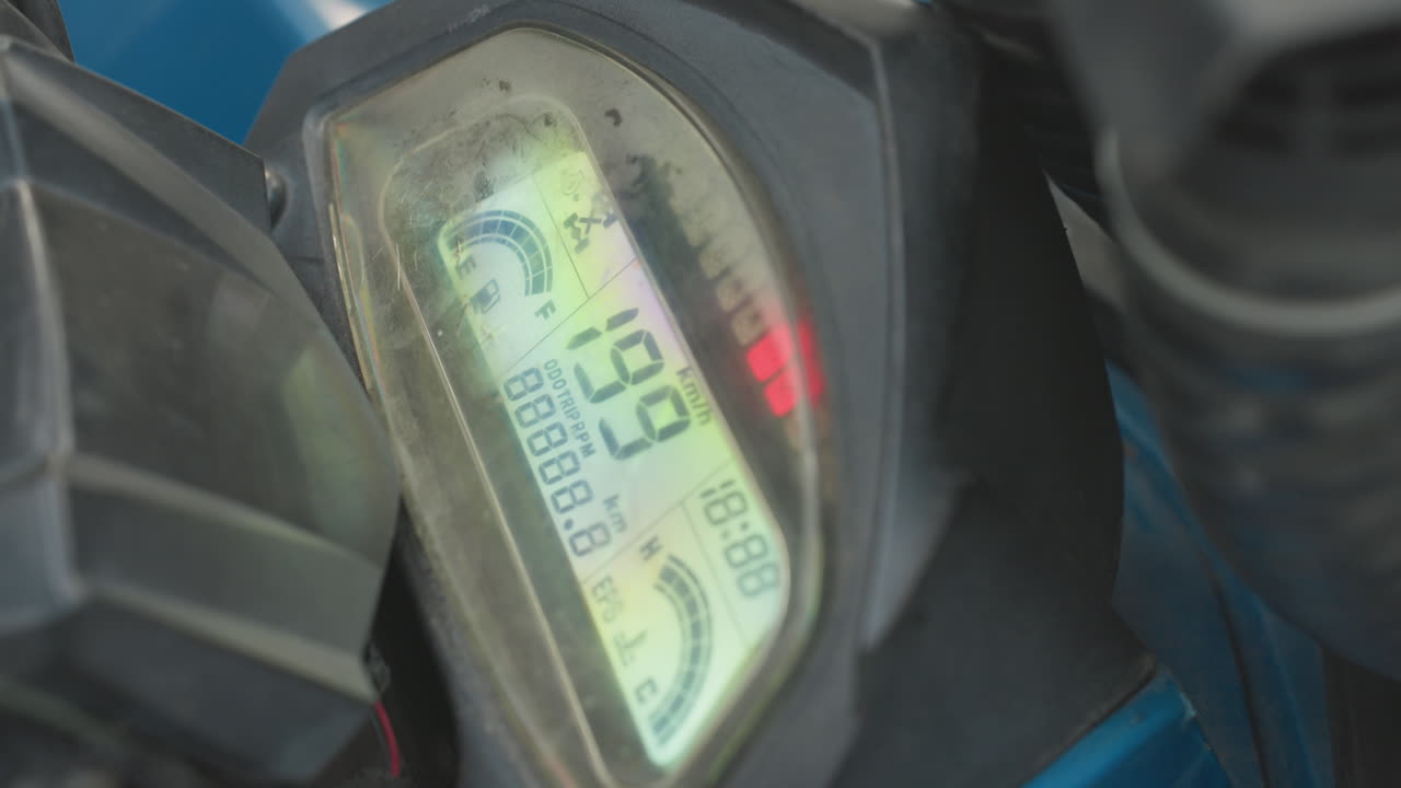 Atv dashboard display showing digital readouts, scratched plastic cover, illuminated lcd odometer and rpm numbers, red warning led glowing, blue bodywork and handlebar cables in frame, macro closeup