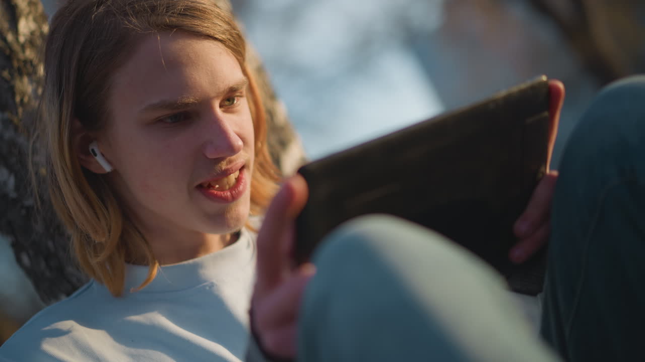 adolescente caucásico, frustrado y concentrado, utilizando una tableta; su expresivo rostro muestra confusión y, después, determinación; toca la pantalla, lleva puestos auriculares, al aire libre al final del día.