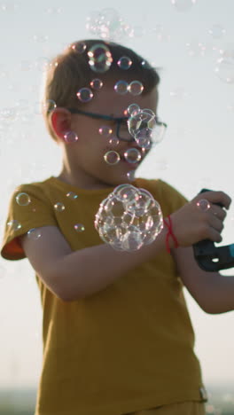 young boy wearing a yellow shirt and shorts, with glasses, stands outdoors, holding a plastic container that releases bubbles into the air. in a bright, open field