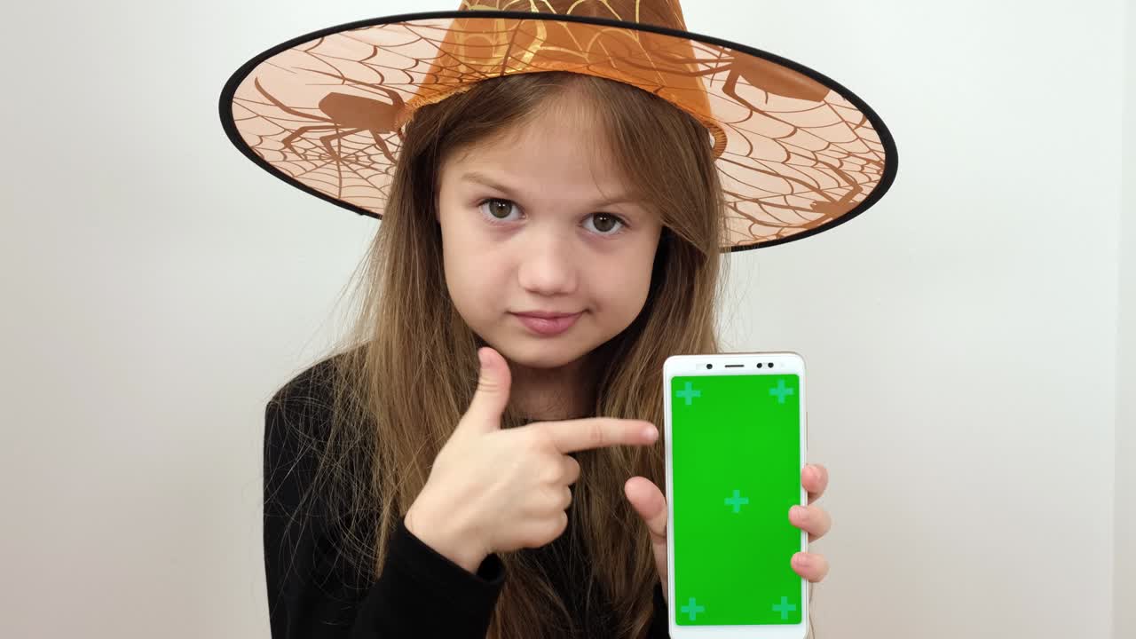 la niña con el disfraz de bruja de halloween muestra el teléfono con una pantalla verde. chromakey pantalla verde teléfono inteligente configurado para la publicidad. niño lindo mirando la cámara. niño adorable con dispositivo digital