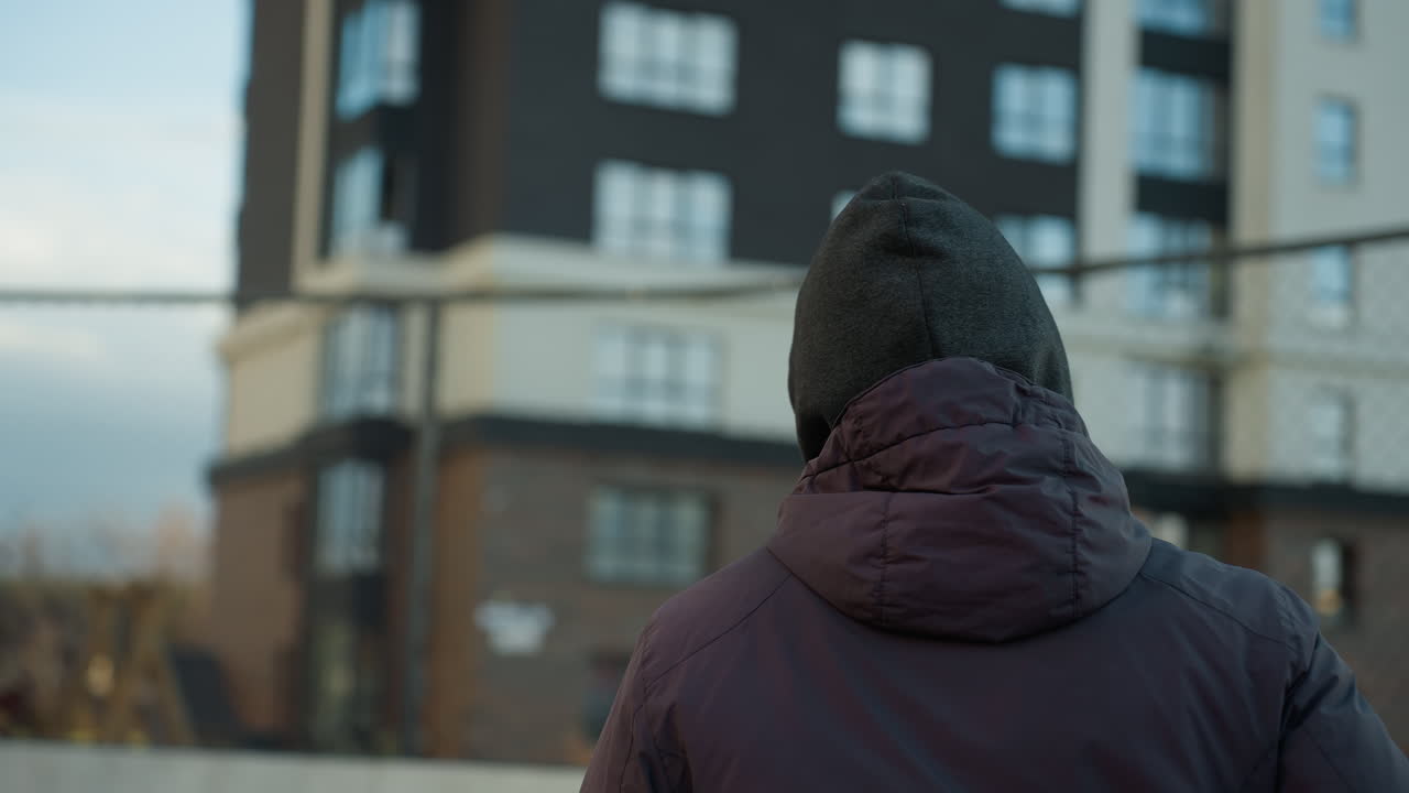 atleta con capucha y gorra corriendo en una arena deportiva con poste de portería en el fondo y vista borrosa de personas caminando, rodeado de arquitectura urbana