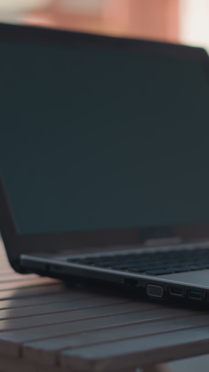 Close-up of person in black coat picking up coffee cup from table with laptop nearby, table setup includes coffee cup and open laptop with blurred background featuring buildings and greener