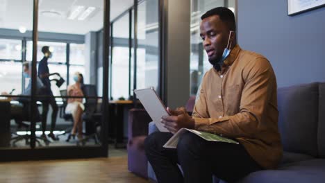 African-american-man-with-lowered-face-mask-having-video-chat-on-digital-tablet-at-modern-office