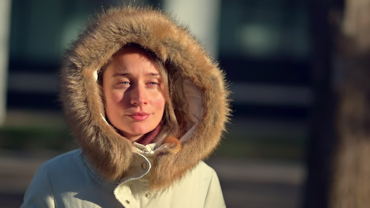 A woman stands outdoors on a sunny winter day wearing a cozy coat with a furry hood. She smiles and enjoys the pleasant weather in a city park, surrounded by trees