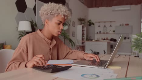 african woman calculates costs using calculator and laptop sit at home desk