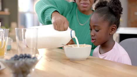 happy african american mother pouring milk on cereal with daughter in kitchen, slow motion