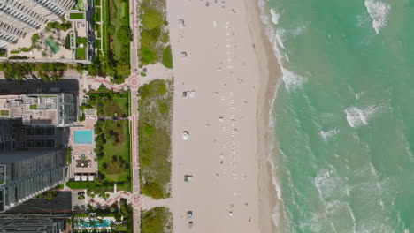 Fly-above-sand-beach-on-sea-coast.-Modern-luxurious-high-rise-hotels-with-swimming-pools-and-palm-trees.-Miami,-USA