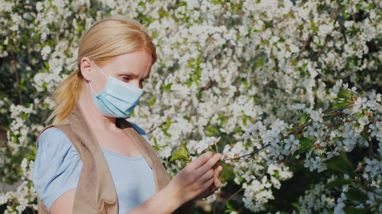 женщина в марлевой повязке рассматривает цветок на фоне цветущего дерева проблемы аллергических заболеваний