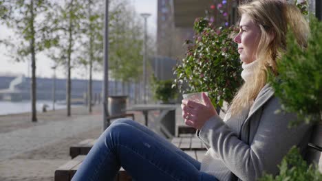 Trendy-young-woman-enjoys-a-cup-of-coffee-in-the-morning-sunlight,-sitting-on-a-bench