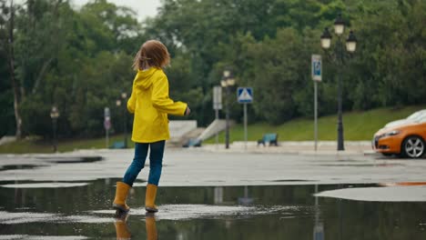 happy blonde teenage girl in a yellow jacket and orange rubber boots jumps in a puddle and splashes water to the sides while walking in the park after the rain