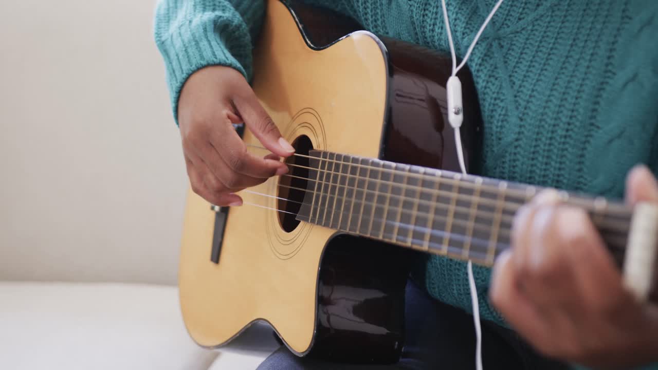 sección media de una mujer afroamericana practicando la guitarra y usando auriculares en casa, cámara lenta
