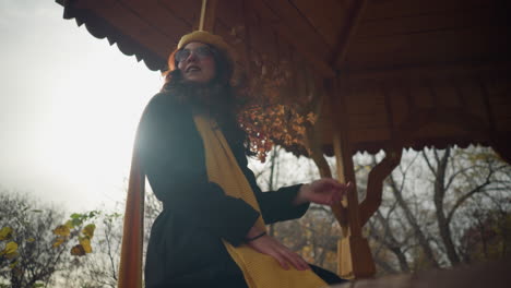 stylish lady seated in a gazebo, playfully stretching her red hair with sunglasses on, yellow muffler draped around, set against a backdrop of autumn trees