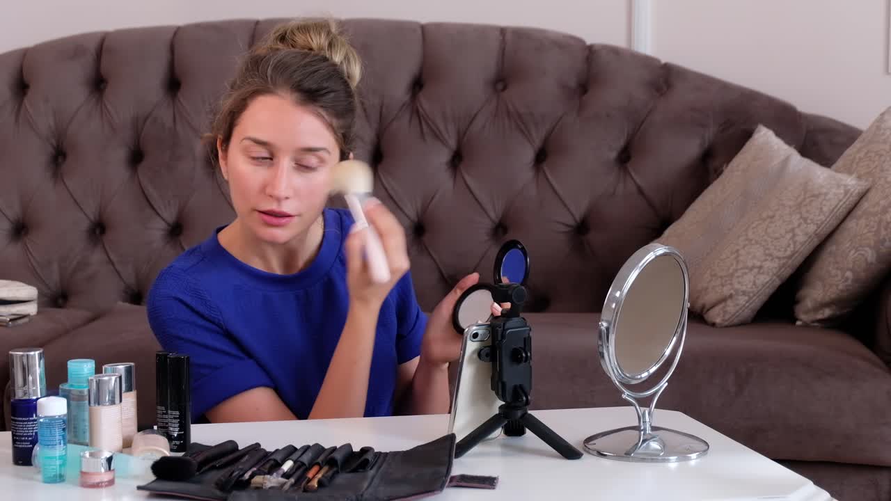 Woman in a blue T-shirt filming herself while doing a make-up tutorial at home