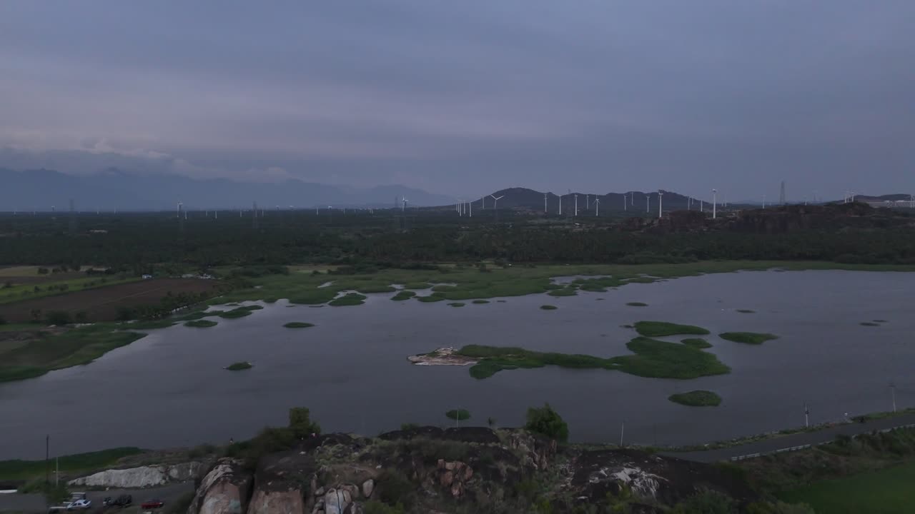 Vista aerea di un paesaggio rurale al crepuscolo. Un lago si trova in primo piano, con un parco eolico visibile sulle colline sullo sfondo. Una catena montuosa distante incornicia la scena sotto un cielo nuvoloso e malinconico.