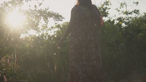 Toma-En-ángulo-Bajo-De-La-Espalda-De-Una-Mujer-Caucásica-Caminando-Por-Un-Campo,-Los-Rayos-Dorados-De-La-Distante-Puesta-De-Sol-Brillando-A-Través-De-Los-Huecos-De-Los-árboles-En-Un-Hermoso-Día-Sin-Preocupaciones