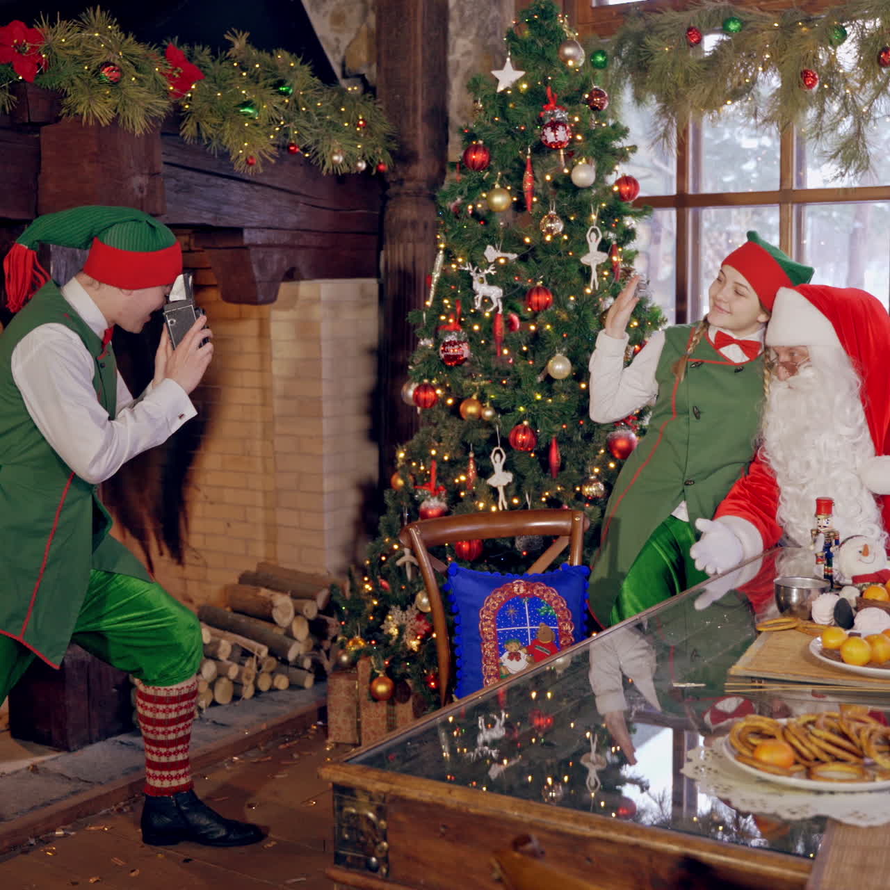Santa Claus together with fairy elves. One elf is taking photo by retro camera. Female elf and Santa photographing on Christmas decorated room.
