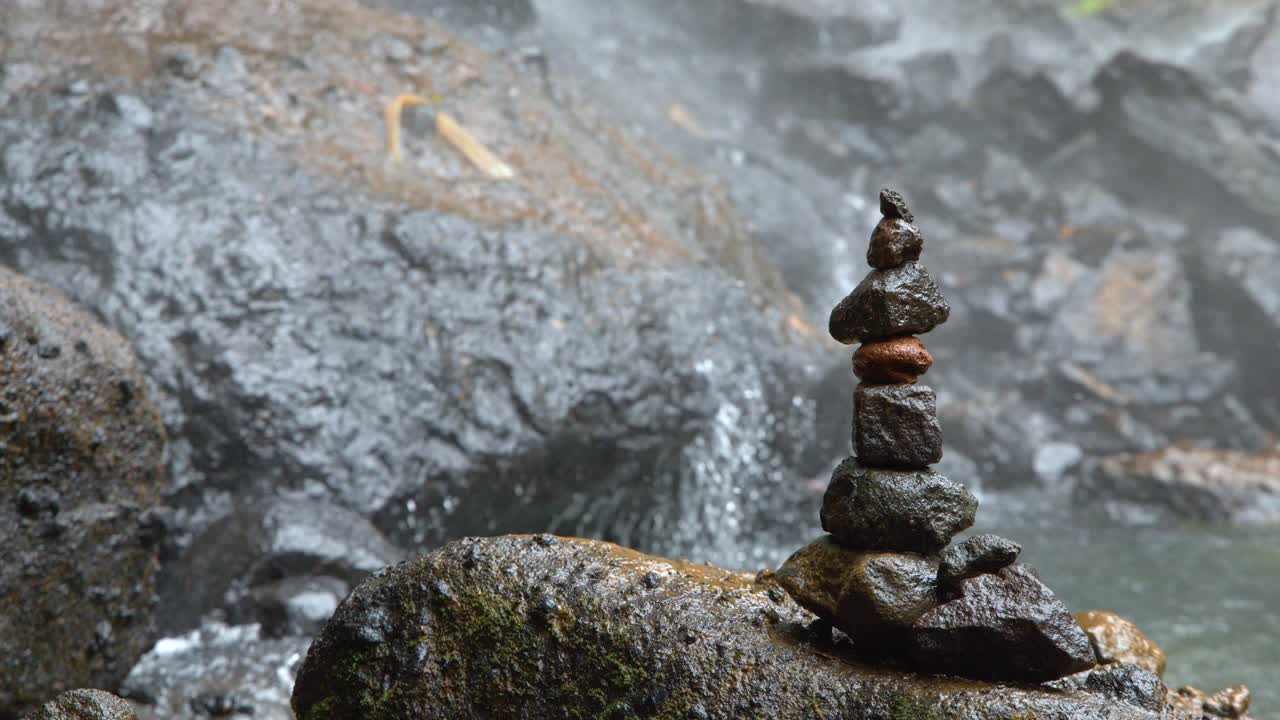 Inquadratura statica media di pietre impilate in equilibrio su massi bagnati vicino a una cascata tropicale nebbiosa.