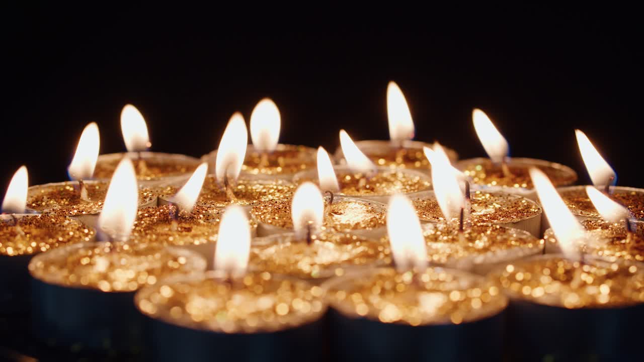 Sfondo suggestivo con candele tealight accese, che brillano di un giallo dorato intenso, per celebrazioni, occasioni religiose, anniversari, feste o in memoria delle vittime di un tragico attacco. Una candela ruota velocemente e lentamente.