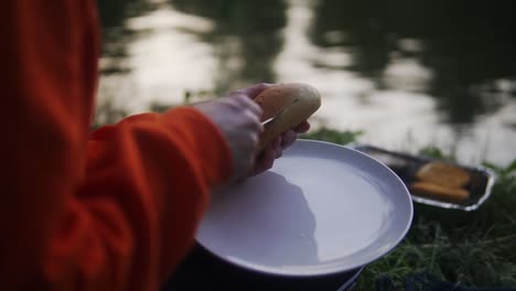 a close up shot of a lady wearing full sleeve dress cutting burger bun while sitting along side of a pond