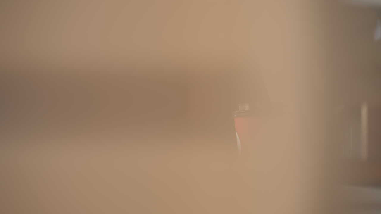 Close-up of coffee cup on table with person sitting, partially obscured by laptop edge, indoor workspace setting featuring blurred background