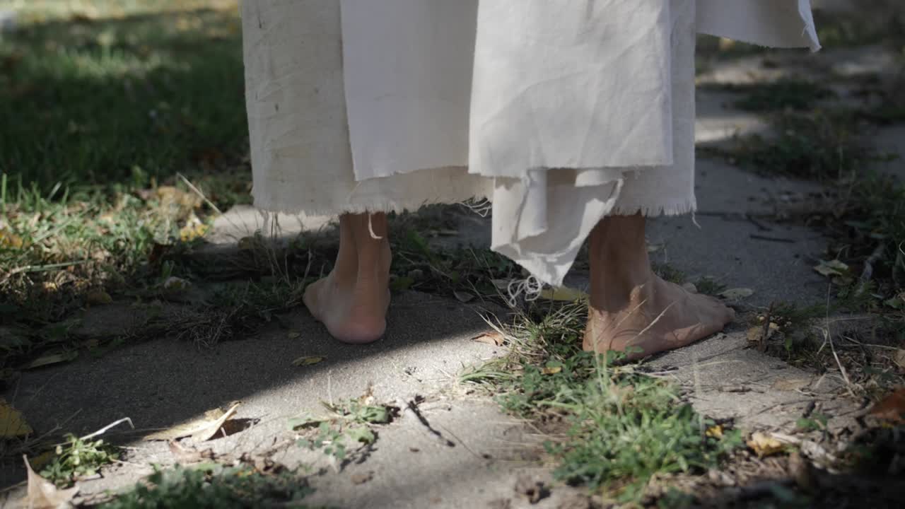 흰색의 너덜너덜한 가운을 입고 예수 그리스도처럼 옷을 입은 남자가 보도에서 슬로우 모션으로 서 있다