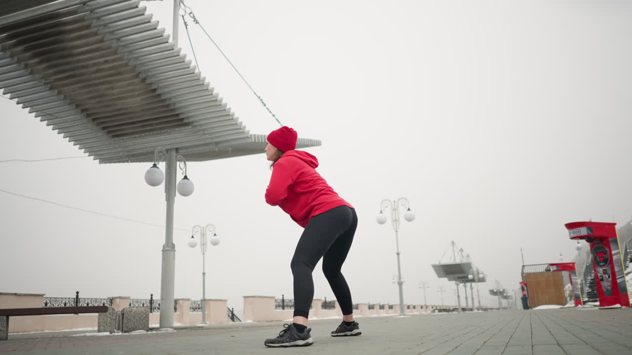 Side view of woman performing side lunge exercise outdoor, she is dressed in red hoodie and black leggings, exercising on a snowy urban promenade with light poles, steps, and a public machine