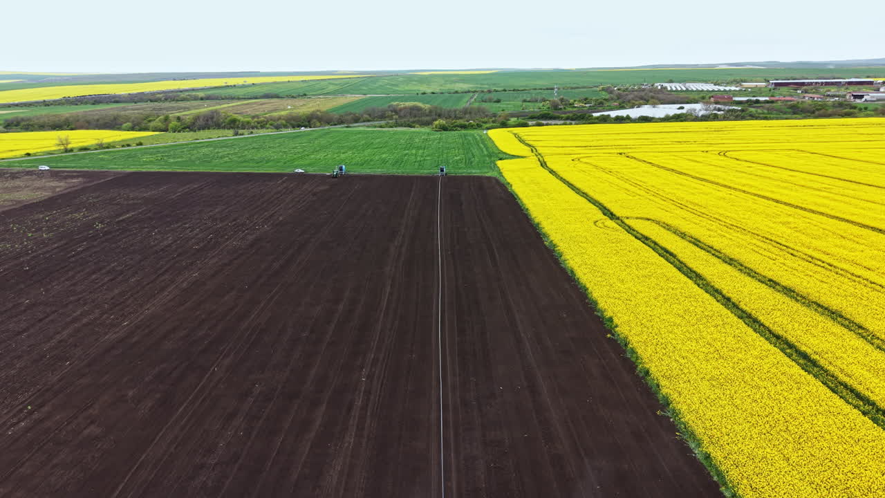 Landwirtschaftliche Geräte bewässern tagsüber Felder in einer ländlichen Gegend mit sichtbaren grünen und gelben Ackerflächen.
