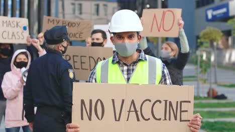 caucasian builder wearing protective mask and helmet holding no vaccine" singboard and looking at the camera in a protest against covid 19"