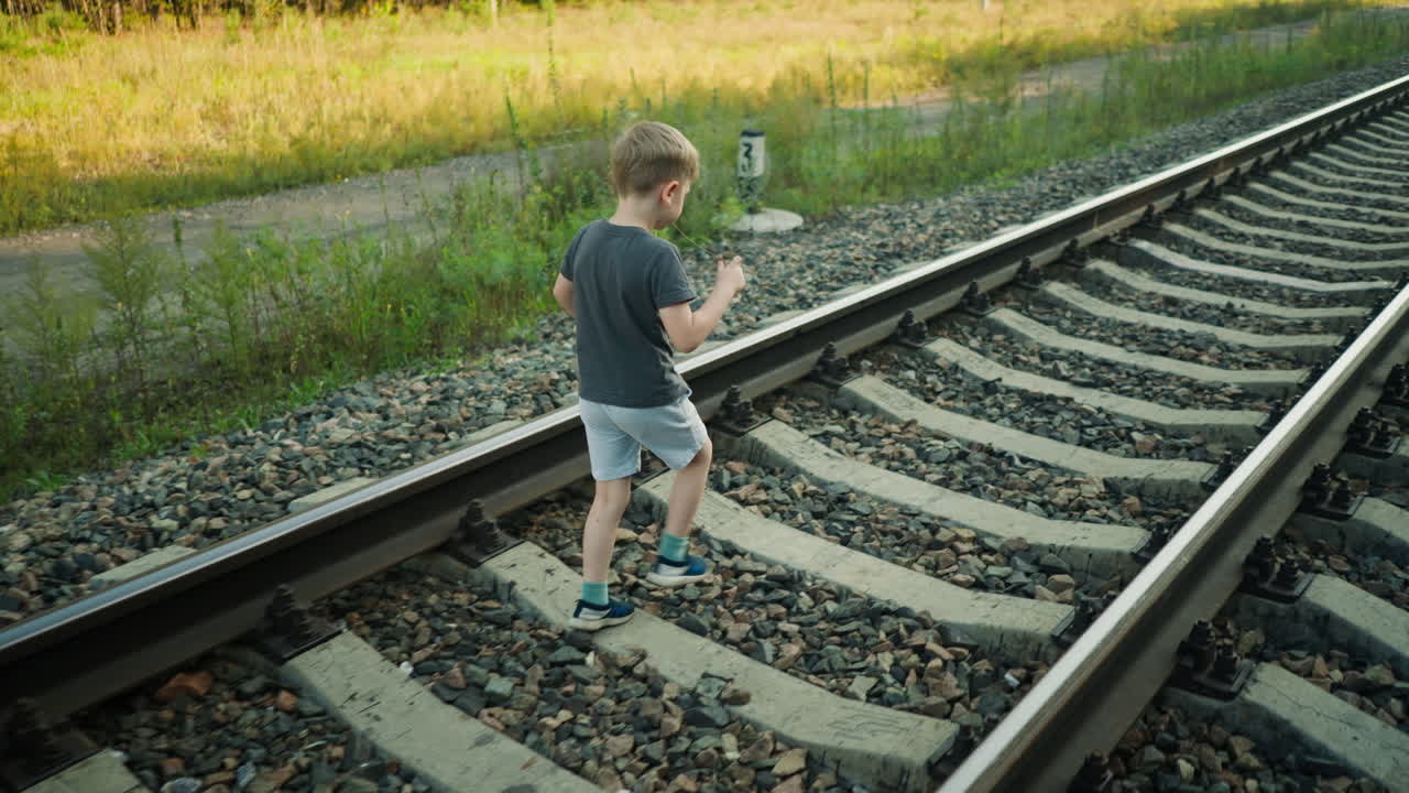 side view of young boy in casual clothes walking alone on rail track surrounded by gravel and grassland, early evening light, quiet outdoor rural setting with roadside and overgrown vegetation