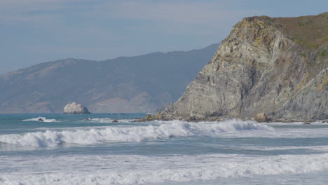 Tiro-Estacionario-De-Olas-Rompiendo-En-El-Lado-De-Un-Acantilado-Con-Montañas-En-El-Fondo-Ubicado-En-La-Playa-De-Big-Sur-California