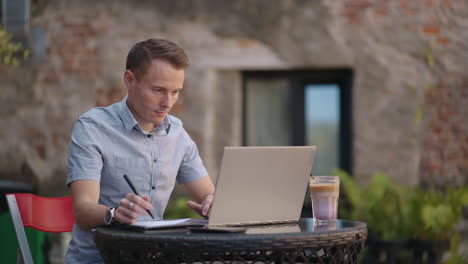 a man sitting in a street cafe works remotely writing a pen in a notebook. work at the laptop. a man works remotely on a laptop