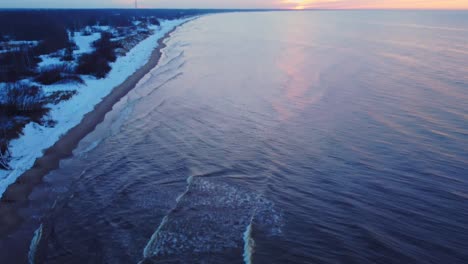 aerial view of beach covered in snow with waves crashing, tilt reveals sunset