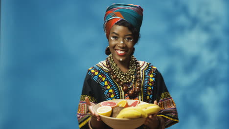 joven afroamericana alegre con turbante y maquillaje sonriendo alegremente a la cámara mientras baila y sostiene un plato con frutas