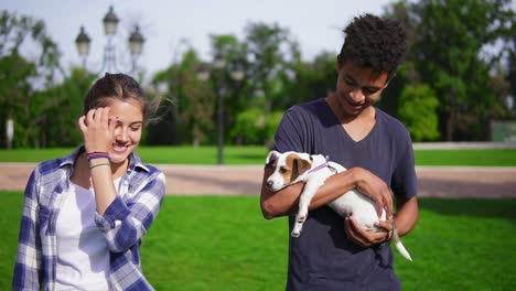 Two-multiracial-friends-walking-in-park-enjoying-the-summer-day.-Young-attractive-african-guy-is-holding-cute-little-jack-russell