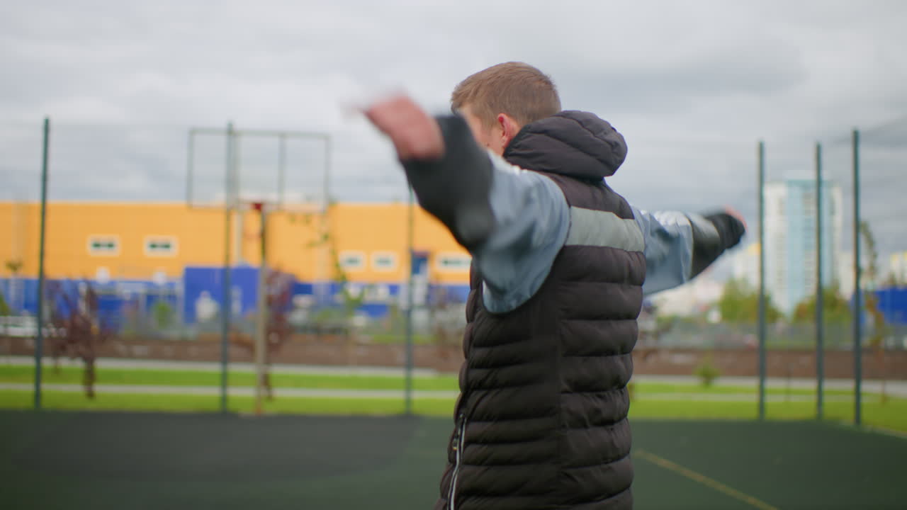 Jovem caucasiano se alongando em campo, usando colete puffer e blusa de moletom com capuz, fazendo rotações lentas com os braços e simulando arremessos, céu nublado com prédios residenciais altos e cerca ao fundo, rotina de aquecimento focada