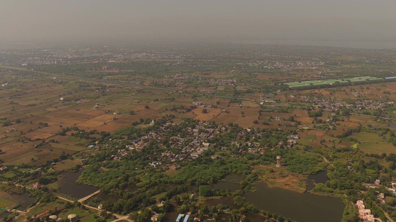 Una ripresa cinematografica con drone ad alta quota che mostra un tradizionale villaggio indiano circondato da alberi verdi e lussureggianti, campi agricoli e piccoli laghi sotto un cielo velato