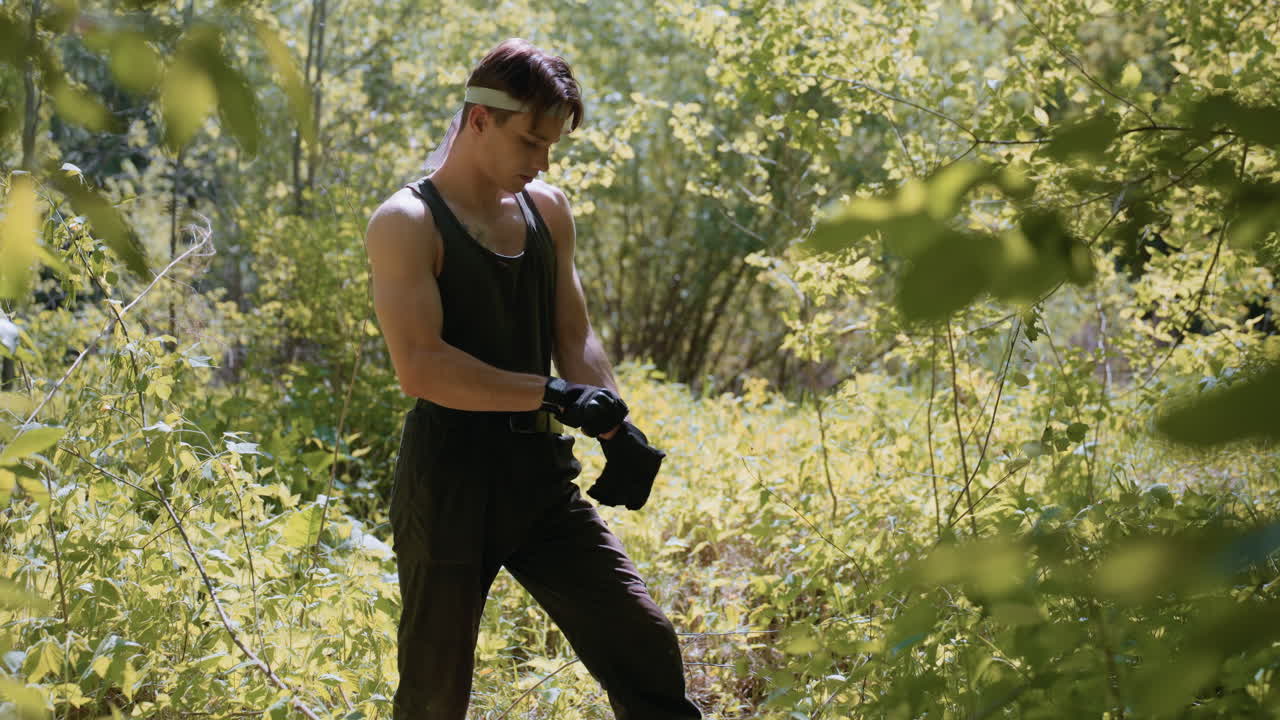 Soldier putting on gloves while standing in slanted stance with one leg forward and other back, adjusting hand strap amid dappled sunlight filtering through dense forest foliage on summer trek
