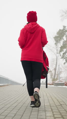 back view of lady in red winter jacket and head warmer holding red backpack walking along fog-covered pathway lined with snowy benches and frosted trees on cold winter day, looking away into distance
