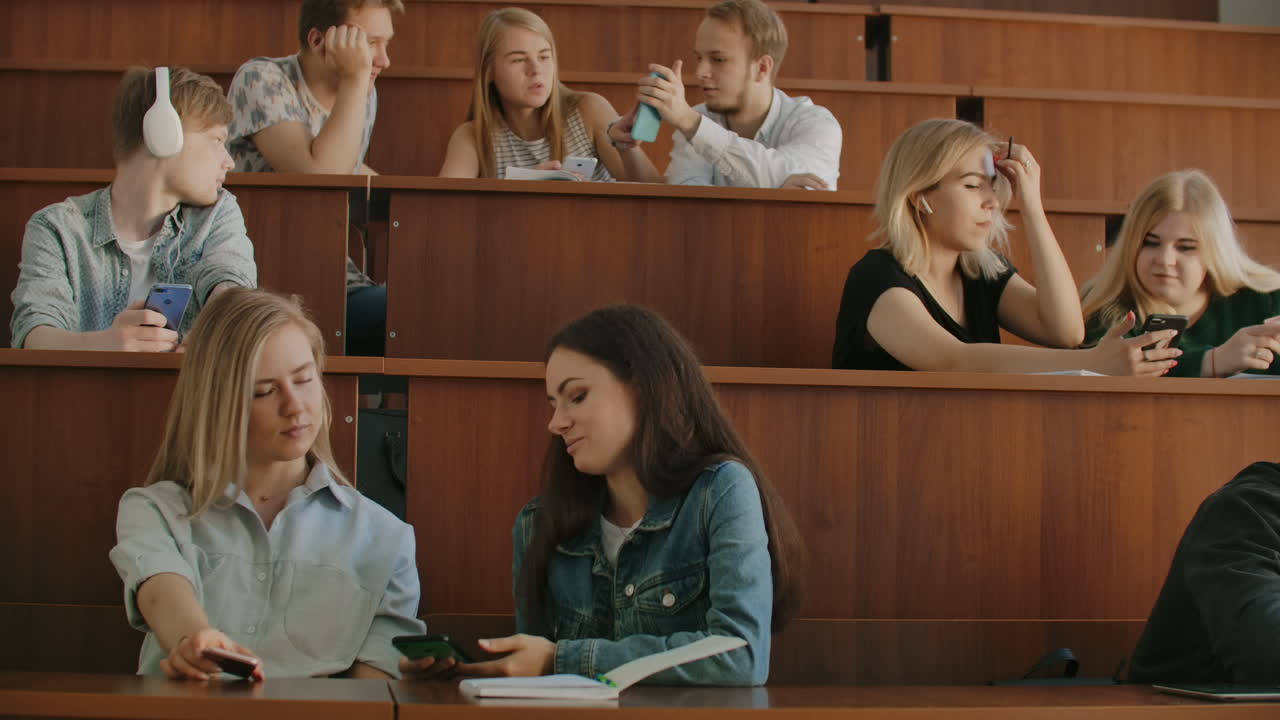 Pretty girls students are using smartphone watching screen talking and laughing sitting at desks at university. Social media internet millennials and education concept