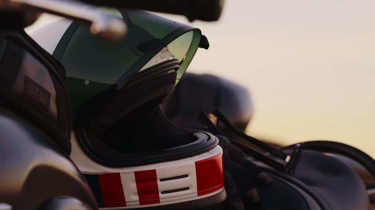 Primo piano statico di un casco da motociclista in stile vintage o retrò con visiera colorata, appoggiato sul manubrio o sul sedile di una moto sotto la luce del sole intensa e velata. L'attenzione è rivolta ai dettagli e allo stile.