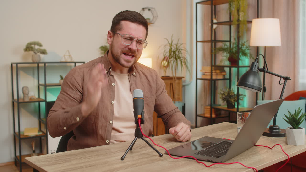 Un giovane influencer caucasico felice che parla al microfono e alla webcam del portatile al tavolo dell'ufficio a casa