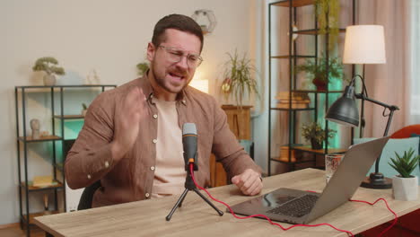 happy young caucasian man influencer speaking into microphone and laptop webcam at home office table