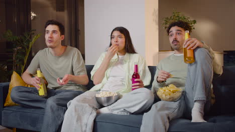 three friends watching interesting movie on television sitting on couch, eating popcorn and chips and drinking soda 3