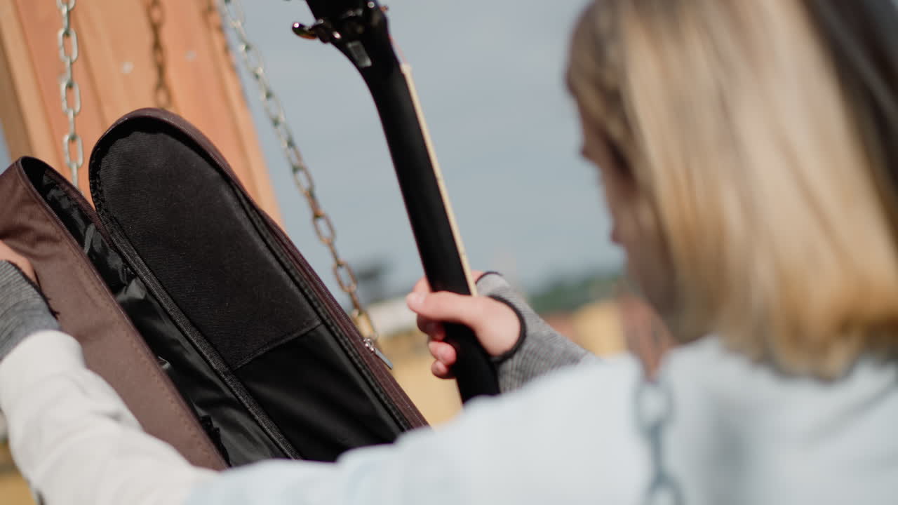 Una donna bianca bionda apre la custodia di una chitarra, solleva uno strumento nero da una borsa imbottita su una banchina del fiume, mani sicure fanno scivolare il manico nella luce, skyline della città sfocato sullo sfondo, musicista che prova per la sessione