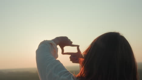 rear view of young woman framing sun with hand gesture, radiant sunlight glowing through fingers, soft lens flare, blurred riverscape horizon, serene golden hour
