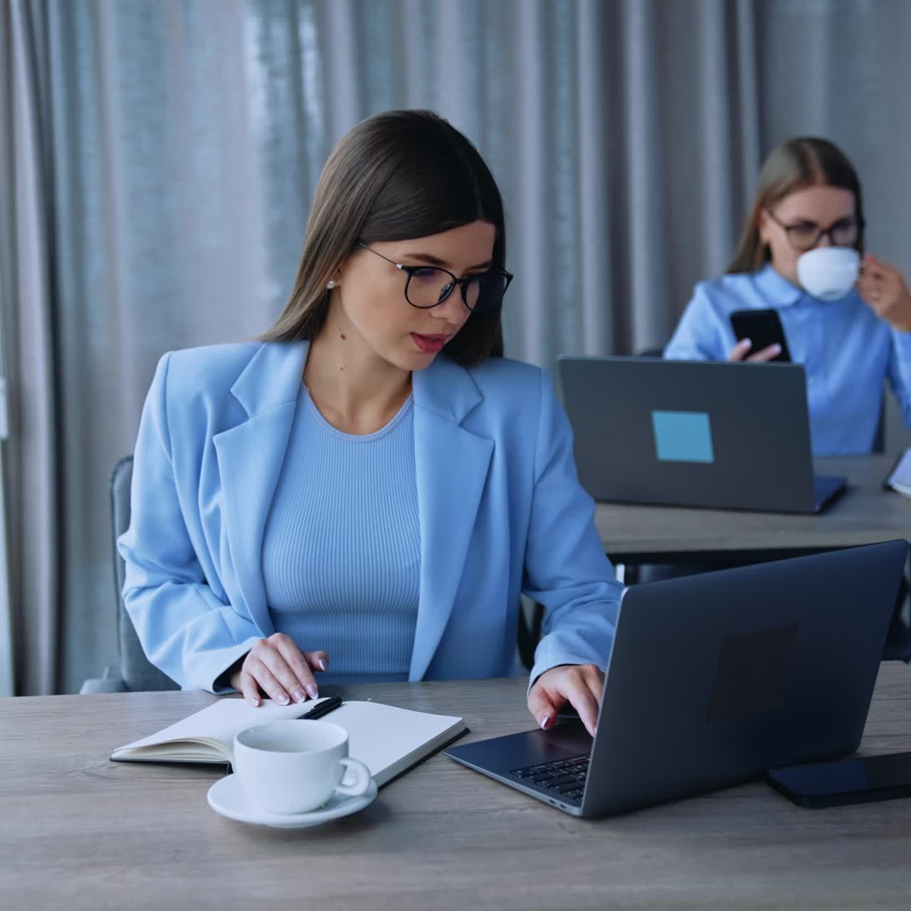Young ladies working in the office on laptops. Brunette woman looks at the screen and Blonde one drinking coffee and looking at phone. Blurred backdrop