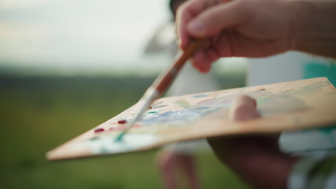 화가의 손이 팔레트를 들고 으로 페인트를 적용하는 클로즈업, 배경에 여성의 흐릿한 형상이 장면에 깊이를 더합니다.