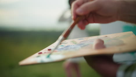 a close-up of a painter's hand holding a palette, applying paint with a brush, while a blurred figure of a woman in the background adds depth to the scene