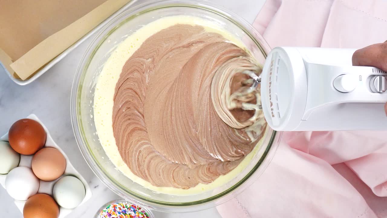 Baking chocolate brownies and using a white electric mixer to mix the batter. Making brownies on a white background surrounded by eggs, pink napkin and a baking pan. In the kitchen making a dessert