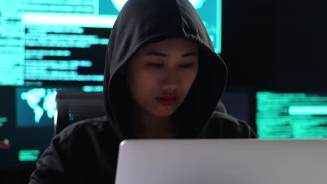 close up of asian female hacker in a hood works on a laptop with maps and data on display screens of multiple monitors in a dark office room